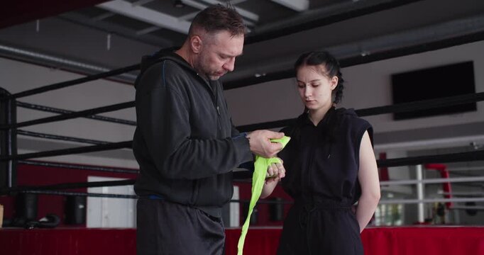 A trainer applies protective wraps to a young female boxer's hands before training, providing protection and support to her wrists and knuckles in preparation for intense boxing training.