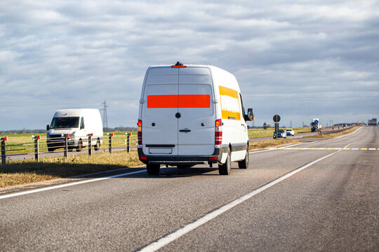 White delivery van traveling highway on cloudy day, rear view of cargo transport vehicle with orange stripe on back doors, journey on asphalt road with power lines in background
