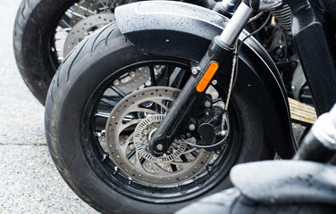 Obraz premium Close-up view of front wheel of motorcycle, showing tire, brake disc, suspension, and fender covered in raindrops, vehicle parked outdoors on asphalt, detail of modern bike engineering