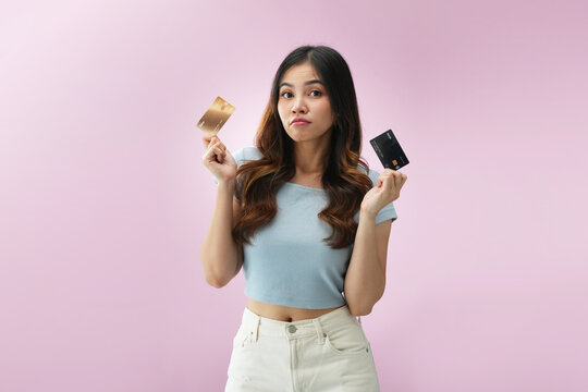 Confused Indonesian woman holding two credit cards with doubtful expression on pink background, illustrating financial decision making, payment comparison, and consumer choice concept.