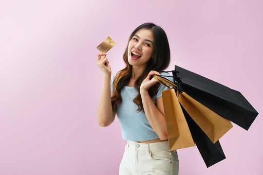 Happy Indonesian woman carrying multiple shopping bags while holding credit card with excited expression on pink background, representing fashion shopping, retail sale, cashback promo, and consumer sp