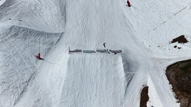 Drone view of snowboarders and skiers performing tricks on big air kickers in Tignes snowpark during sunny spring day in the French Alps. Freestyle snowboard and ski session on big air jumps in alps