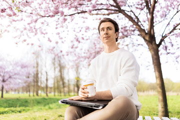 Obraz premium A male freelancer takes a relaxing coffee break from remote work outdoors, sitting on a park bench with his laptop under blooming pink cherry blossom trees in spring.