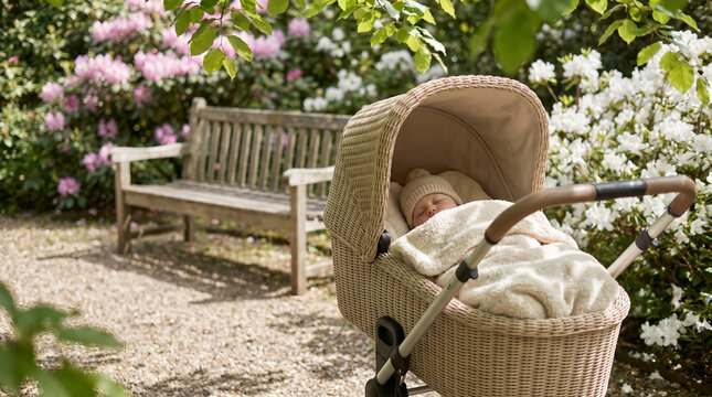 Newborn baby sleeping peacefully in wicker bassinet stroller in blooming garden with wooden bench