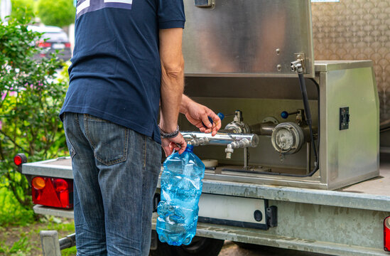 Man pouring drinking water from a mobile cistern into a large, blue plastic bottle, metal tap of a water tanker on a trailer, back view, daylight, close-up on hands and water dispenser.
