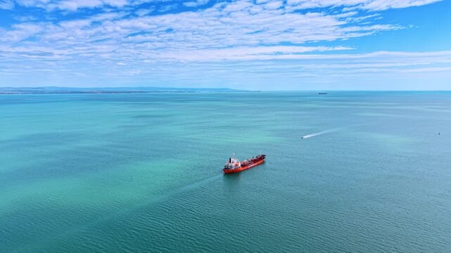 Industrial oil tanker vessel cruising turquoise ocean water. High angle drone shot of a red tanker ship moving through calm turquoise sea with distant coast.