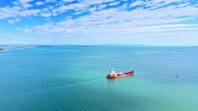 Red oil tanker ship sailing in turquoise sea. Aerial shot of an orange and red tanker vessel cruising through bright turquoise water leaving a white wake.