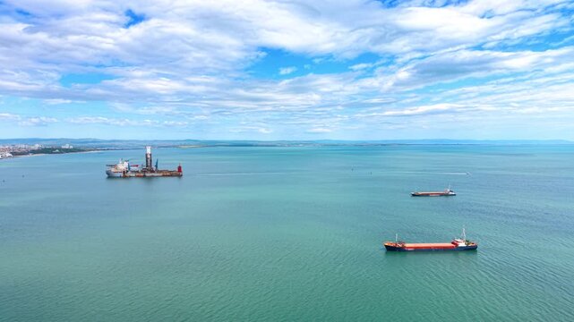 Wide aerial view of offshore drillship and cargo vessels at sea. High angle panoramic shot showing an industrial drilling ship and two cargo boats floating in open sea near coast.