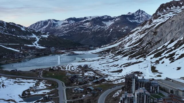 Evening drone view of Tignes 2100 in the French Alps, showing illuminated mountain resort buildings and hotels surrounded by dramatic alpine peaks under twilight sky atmosphere