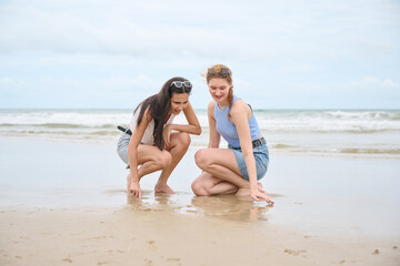 Fototapeta premium Young women are squatting on a wet sandy beach, looking down at something they are interacting with in the sand, with the ocean and a cloudy sky in the background