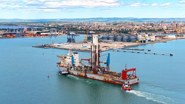 Marine specialized drilling ship navigating near industrial port terminal. Technical aerial drone view of offshore well intervention vessel accompanied by tugboat at commercial seaport.