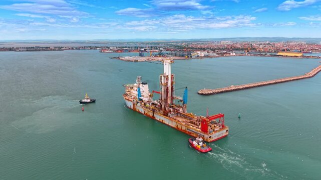 Industrial offshore drillship assisted by tugboats near sea port. Technical aerial shot of specialized drilling vessel with central tower and tugboats navigating a harbor basin.