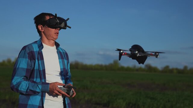 Man flying FPV drone with goggles outdoors. Pilot controls drone with remote controller in green field. FPV goggles provide first-person view for drone pilot. Quadcopter hovers near drone operator.
