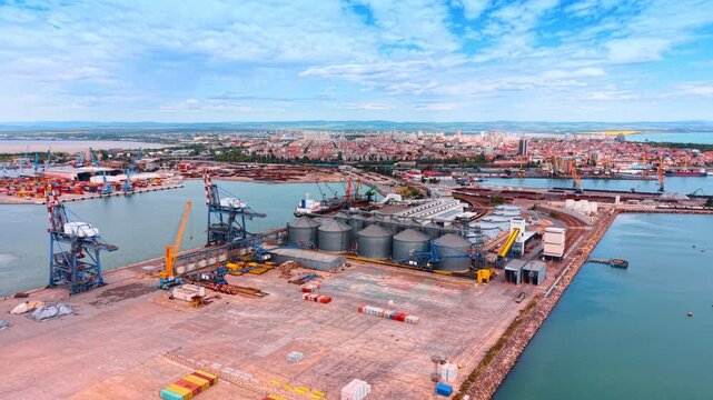 Wide aerial view of industrial harbor docks and cargo terminal. Panoramic drone perspective of sea port docks with grain silos, container cranes, and urban coastline.