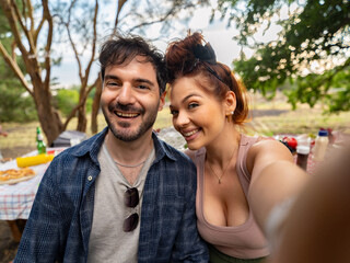 Fototapeta premium Happy young couple smiling while taking selfie during outdoor picnic in park