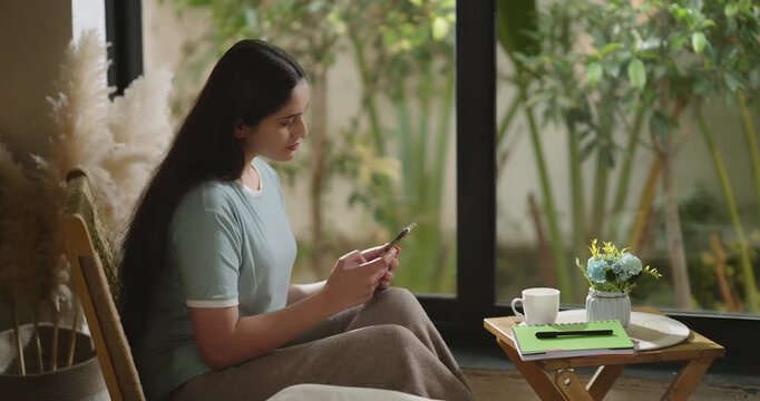 Indian young adult gen z relax female girl sitting on chair use mobile do virtual chat take job break room happy woman hold phone talk speak video call share new gossip enjoy day spend time home place