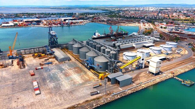 Aerial view of grain terminal and silos in sea port. Top view of large industrial grain elevator complex with metal silos, railway tracks, and cargo docks at sea port.