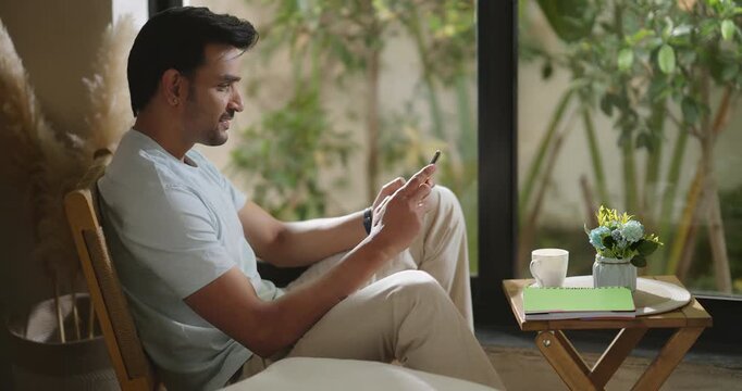 Indian young adult gen z relax male boy sitting on chair use mobile do virtual chat take job break room happy man guy hold phone talk speak video call share new gossip enjoy day spend time home place