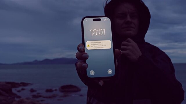 Woman in Hooded Jacket Holding Smartphone With Hurricane Warning Near Rocky Coastline, Extreme Weather Awareness and Personal Safety