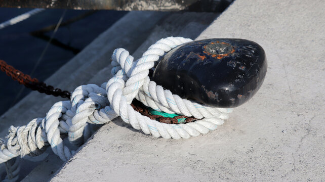 Close-up of a thick, braided white nautical rope securely fastened around a weathered black metal mooring bollard on a concrete pier, with a rusty chain in the background. Symbolizing maritime securit