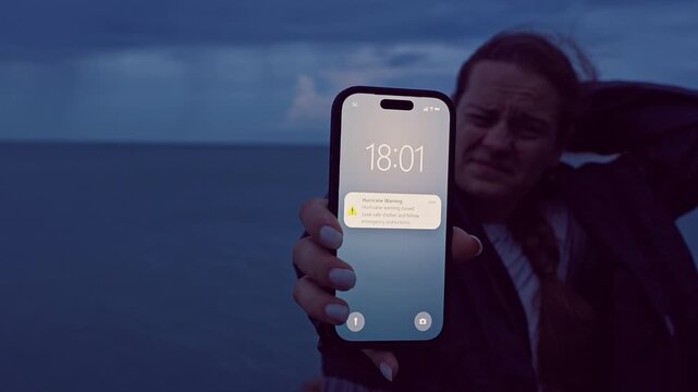 Woman Holding Smartphone With Hurricane Warning While Struggling Against Strong Coastal Wind at Dusk, Emergency Alert and Severe Weather Survival