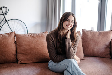 Obraz premium Smiling Young Woman Sitting On Couch In Cozy Living Room At Home Wearing Casual Sweater