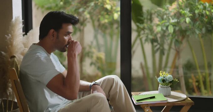 Indian calm young adult gen z relax man sitting on chair pick up cup drink coffee enjoy day room. Male boy hold pen book writing new work notes plan ideas spend time take casual job break home place