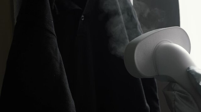 A close-up shot of white steam coming out of the nozzle of a vertical garment steamer. Hot humid air is directed towards the fabric of a dark polo shirt