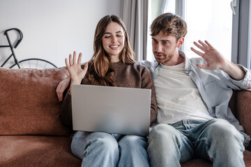 Young Couple Waving During Video Call On Laptop While Sitting Together On Couch At Home