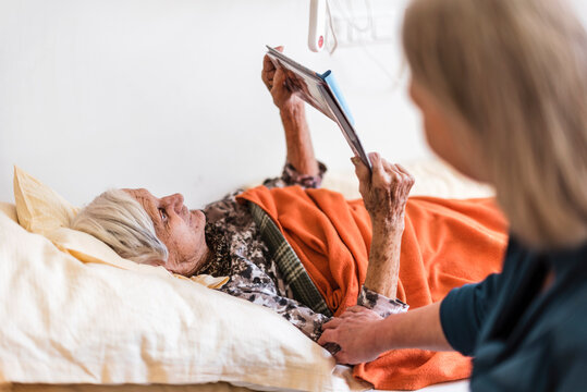Woman taking care of old woman lying in bed reading book