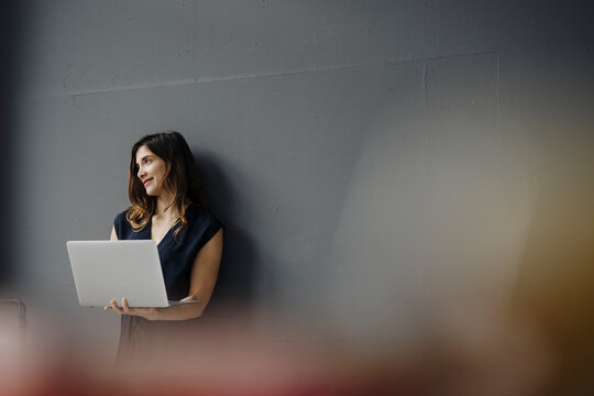 Portrait of smiling young woman with laptop leaning against wall looking at distance