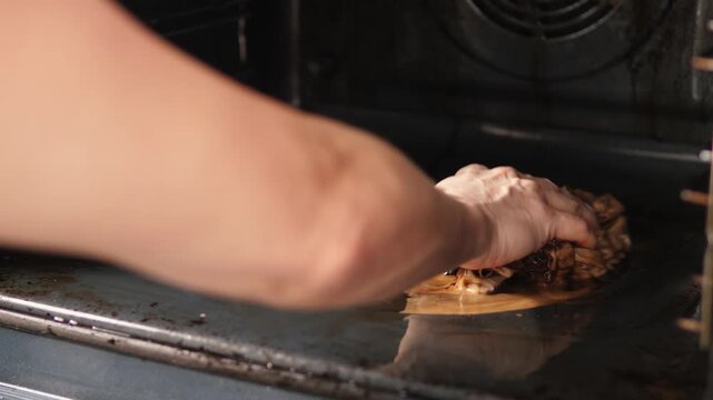 Woman's hand cleaning a very dirty oven bottom from burnt grease and carbon deposits with a rag. The process of deep cleaning kitchen appliances and removing stubborn stains