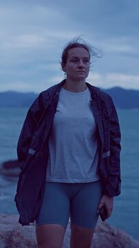 Vertical video. Slow Zoom Shot of Woman Walking Through Strong Coastal Wind Near Stormy Ocean at Blue Hour, Resilience and Survival During Extreme Weather