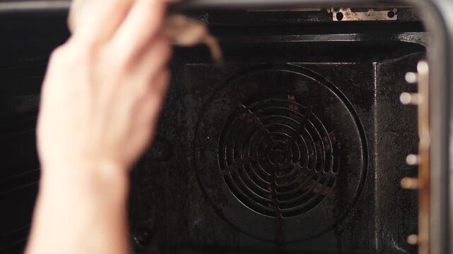 Close-up of a dirty oven interior with a convection fan being cleaned by a woman. Female hand wiping the upper corners and walls with a rag to remove stubborn grease and soot