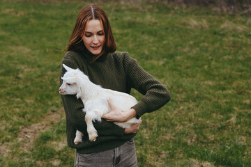Fototapeta premium Lifestyle scene of a woman in a knitted green sweater holding a goat kid in a field, meadow and nature backdrop, warm light, calm affectionate moment outdoors.