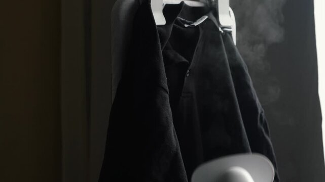 A hand in a heat-resistant glove steams a black shirt on a hanger using a vertical garment steamer. A jet of hot steam effectively removes wrinkles from the fabric