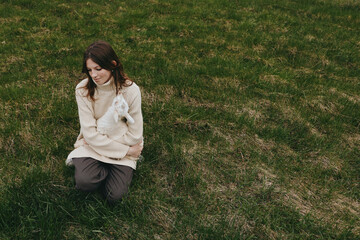 Fototapeta premium A casual lifestyle scene shows a young woman kneeling on grassy ground, cuddling a small white animal, cozy sweater, tranquil moment outdoors, conveying tenderness and connection with pets.