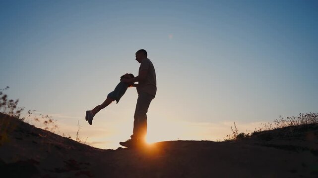 A man carries a boy while standing on a hill. The sun sets in the background, casting silhouettes of the man and boy against the colorful sky.
