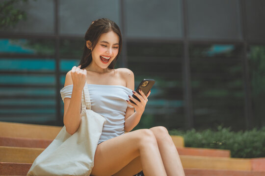 Excited young Asian woman celebrating good news on smartphone while sitting outdoors on steps cheerful female looking at mobile phone screen with fist pump success win and digital consumer lifestyle