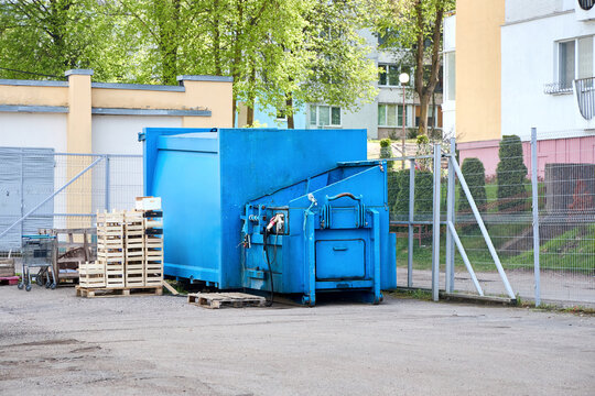 Large blue industrial waste compactor sits on asphalt near wooden pallets and crates. Metal fence and residential building with trees appear in background. Heavy machinery used for urban refuse.