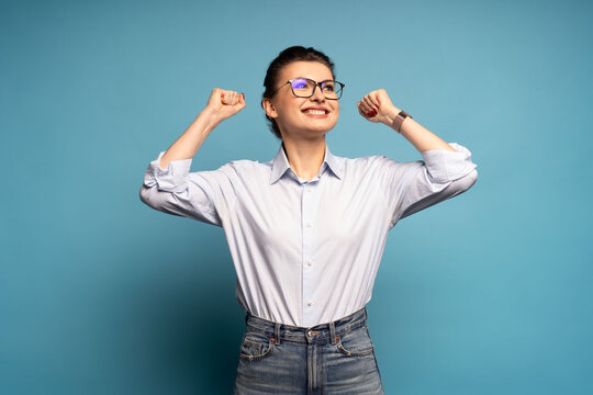 Extremely happy woman standing with raised clenched fists, winning lottery, celebrating her success isolated on blue
