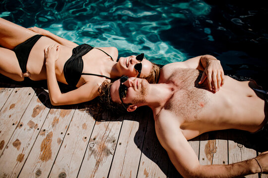 Young hot couple resting together. Side view of cheerful man and woman lying at swimming pool and enjoying relax rest time. Sunbathing after swimming.