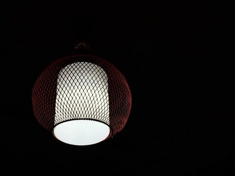 A lit pendant lamp with a bright white cylindrical diffuser surrounded by a reddish woven mesh cage shroud against a solid pitch-black background. An abstract geometric form