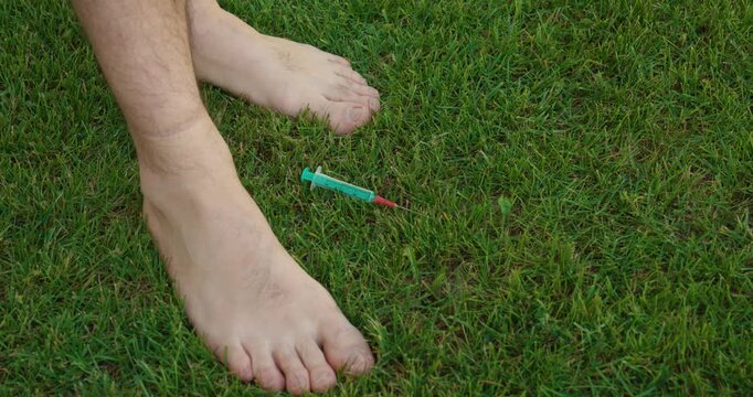 Close-up of bare feet standing on grass near a discarded syringe, showing a dangerous health hazard, risk, and unsafe outdoor environment.