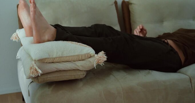 Person resting on a couch with legs elevated on pillows, suggesting dizziness, low blood pressure, circulation problems, fatigue, or recovery at home.