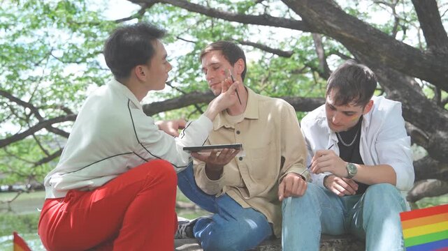 Pride and LGBTQ concept. LGBTQ men sit having a picnic, painting rainbow flags on their friends' faces and smiling happily together in the park.