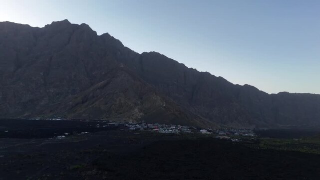 Aerial wide shot of the dramatic Bordeira cliff wall and the remote Ch&atilde; das Caldeiras village situated on black volcanic lava fields at dusk, Fogo Island, Cabo Verde