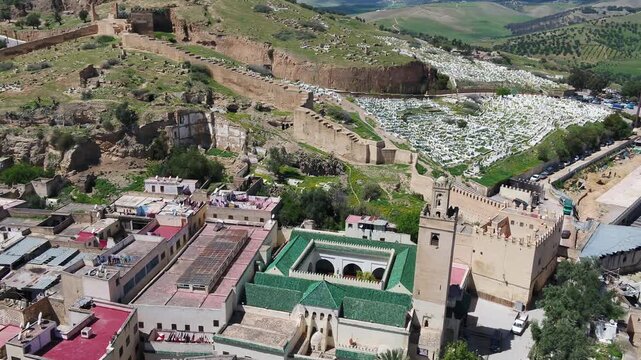 Aerial drone view of historic fortress walls and traditional mosque beneath green hills in Fez Morocco ideal for travel documentary projects