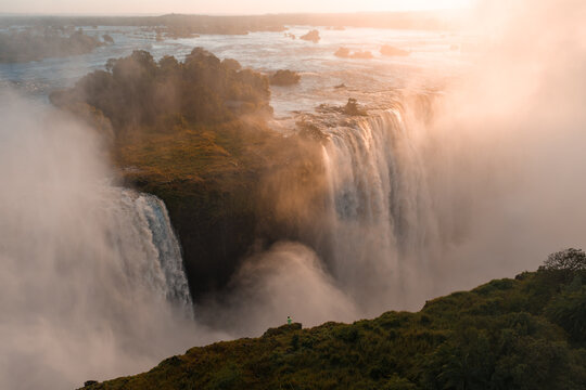 Aerial view shows Victoria Falls at sunset as water plunges into Batoka Gorge, mist rising, Zambezi River spreading over rocky islands, golden light and long shadows.