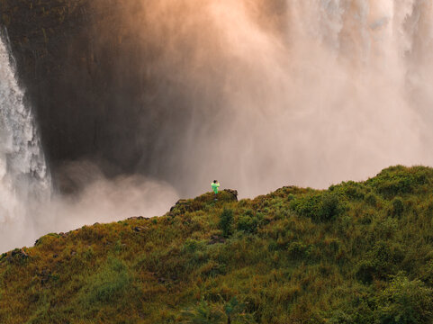 Aerial view shows a person in a bright green jacket on a grassy cliff at Victoria Falls, Zimbabwe, as mist rises and warm sunset light glows through the cascading water.